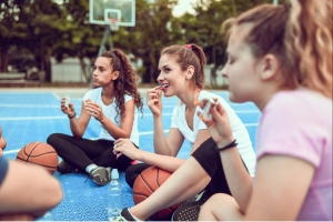 female teen athletes having a snack sitting on basketball court
